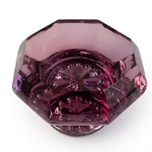 May include: A translucent purple glass bowl with an octagonal rim and a faceted exterior. The interior base features a starburst pattern radiating from the center, while the thick glass walls create a deep violet hue with light reflections.