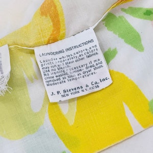 May include: A white fabric with yellow, green, and pink abstract floral designs. A white tag with black text reads "Laundering Instructions Launder whites, colours, and prints separately, as colours may run or stain other fabrics. Use no bleach. No ironing necessary if machine washed, tumble dried, and removed as soon as dryer stops. Moderate temperatures. J. P. Stevens & Co., Inc. NEW YORK N.Y. 10036"