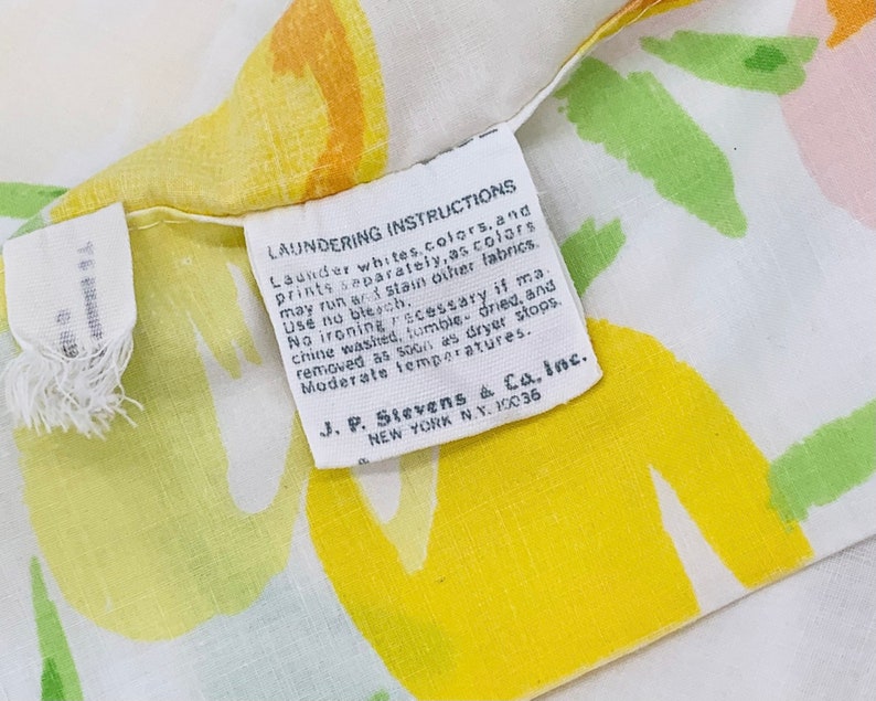 May include: A white fabric with yellow, green, and pink abstract floral designs. A white tag with black text reads "Laundering Instructions Launder whites, colours, and prints separately, as colours may run or stain other fabrics. Use no bleach. No ironing necessary if machine washed, tumble dried, and removed as soon as dryer stops. Moderate temperatures. J. P. Stevens & Co., Inc. NEW YORK N.Y. 10036"