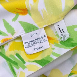 May include: Twin size bed sheet with a white background and a floral pattern in shades of yellow, green, and pink. The sheet has a label that reads "TWIN NO IRON PERCALE 50% POLYESTER 50% COTTON/COTON MADE IN USA. RN-CA-14921 Utica&reg;"