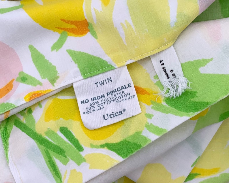 May include: Twin size bed sheet with a white background and a floral pattern in shades of yellow, green, and pink. The sheet has a label that reads "TWIN NO IRON PERCALE 50% POLYESTER 50% COTTON/COTON MADE IN USA. RN-CA-14921 Utica&reg;"