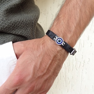 Könnte beinhalten: Ein schwarzes Lederarmband mit einem silbernen bösen Blick-Anhänger. Der Anhänger hat ein blaues Auge mit einer weißen Pupille und einem silbernen Rand.