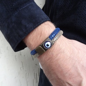 May include: Two braided bracelets, one blue and one beige, are displayed. Both have metal clasps. The blue bracelet features a clasp with an eye design. The bracelets are worn on a wrist.