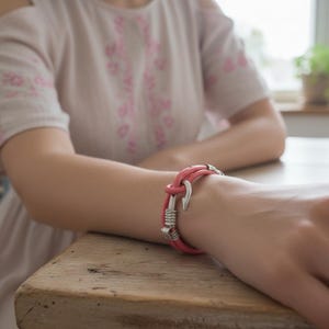 May include: A close-up of a pink leather bracelet with a silver hook clasp. The bracelet is wrapped around a wrist, and the person is sitting at a wooden table. The person is wearing a light pink dress.
