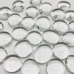 May include: A collection of clear glass stones, also known as marbles or pebbles, with a slightly cloudy or frosted appearance. The stones are smooth and round, and they are scattered on a white surface.