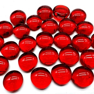 May include: A collection of red glass marbles, scattered on a white surface. The marbles are smooth and round, with a slight shine.