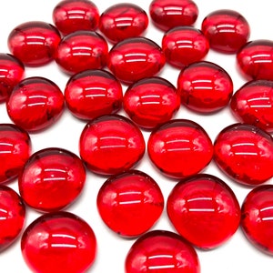May include: A large group of red glass marbles, scattered on a white surface. The marbles are smooth and shiny, with a slight variation in size and shape.