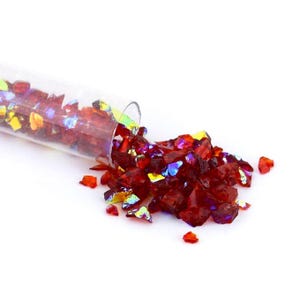 May include: A clear glass tube tilted to spill small, irregular fragments of deep red glass onto a surface. The glass pieces feature an iridescent, multi-colored finish that reflects light with hints of yellow, blue, and purple hues.
