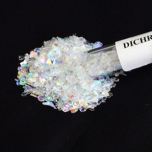 May include: A pile of small, translucent, iridescent flakes with a dichroic finish spilling from a clear glass vial. The flakes reflect various colors including pink, blue, and yellow. A white label on the vial shows the partial text DICHR.