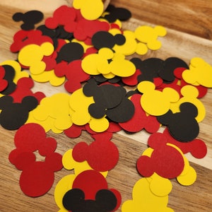 May include: A scattering of red, yellow and black Mickey Mouse head confetti. The confetti is on a wooden surface.