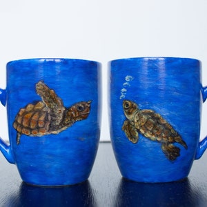 Puede incluir: Dos tazas de cerámica azules con un diseño de tortuga marina pintado a mano. Las tortugas son marrones y tostadas, nadando en un océano azul. Una taza muestra burbujas. Las tazas tienen asas curvas.