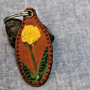 May include: A brown leather keychain with a yellow dandelion design. The keychain has a metal ring and a stitched border. The dandelion has a yellow embroidered flower head, green leaves, and a brown stem.