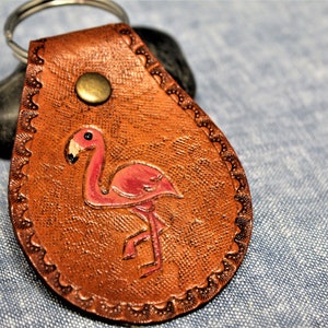 May include: A brown leather keychain with a pink flamingo design. The keychain has a metal ring and a decorative edge. The flamingo is painted with pink and black accents. The keychain is a unique accessory.