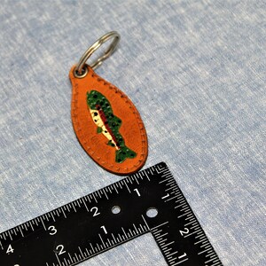 May include: An orange leather keychain with a painted fish design. The fish is green, white, and red. The keychain is oval-shaped and has a silver metal ring attached. The keychain is approximately 3 inches long.