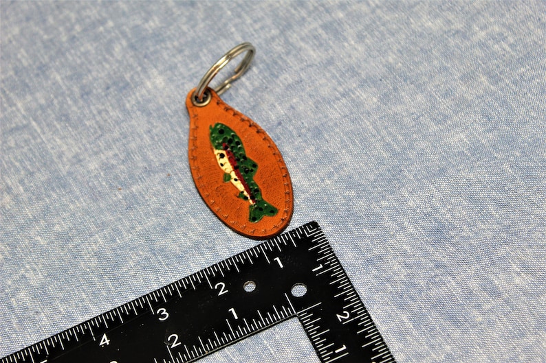 May include: An orange leather keychain with a painted fish design. The fish is green, white, and red. The keychain is oval-shaped and has a silver metal ring attached. The keychain is approximately 3 inches long.