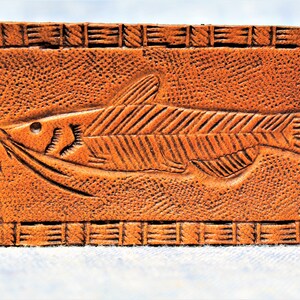 Puede incluir: Un llavero de cuero marr&oacute;n con un dise&ntilde;o de pez tallado. La etiqueta rectangular presenta un relieve detallado de un pez con bigotes y aletas, rodeado por un borde texturizado. Un anillo de metal est&aacute; adjunto para llaves.