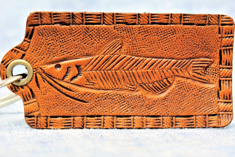 Puede incluir: Un llavero de cuero marr&oacute;n con un dise&ntilde;o de pez tallado. La etiqueta rectangular presenta un relieve detallado de un pez con bigotes y aletas, rodeado por un borde texturizado. Un anillo de metal est&aacute; adjunto para llaves.