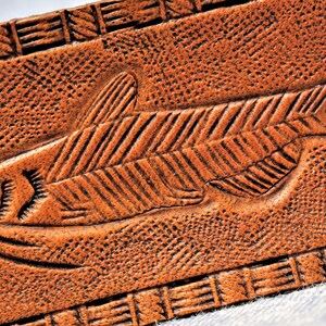 Puede incluir: Primer plano de una secci&oacute;n de cintur&oacute;n de cuero marr&oacute;n con un dise&ntilde;o de pez tallado. El pez est&aacute; detallado con l&iacute;neas grabadas y un peque&ntilde;o ojo. El cuero tiene una superficie texturizada y un borde decorativo.