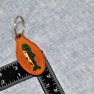 May include: A leather keychain with a fish design. The keychain is oval-shaped and tan with a painted fish in green, red, and white. A silver key ring is attached to the top. The background is a light blue fabric.