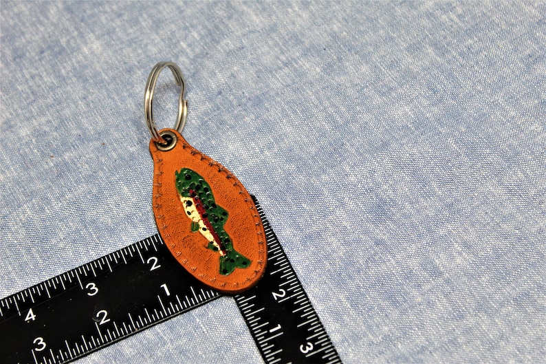 May include: A leather keychain with a fish design. The keychain is oval-shaped and tan with a painted fish in green, red, and white. A silver key ring is attached to the top. The background is a light blue fabric.