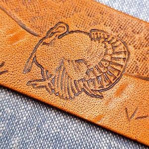 May include: Close-up of a brown leather item with a detailed embossed turkey design. The turkey is centered on the leather, with intricate feather and body details. The leather has a textured surface.
