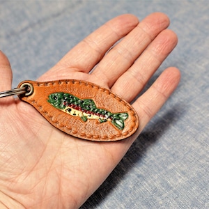 May include: A brown leather keychain with a painted fish design. The fish is green, red, and white, and the keychain has a metal ring. The leather is oval-shaped and stitched around the edges.