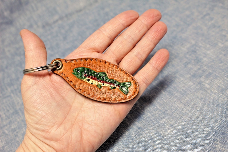 May include: A brown leather keychain with a painted fish design. The fish is green, red, and white, and the keychain has a metal ring. The leather is oval-shaped and stitched around the edges.