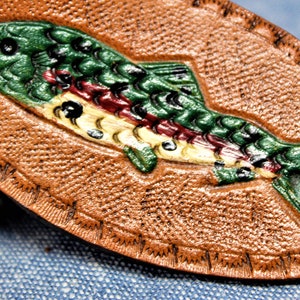 May include: A brown leather oval with a detailed, painted trout design. The fish is green, red, and yellow, with black accents. The leather has a textured surface and a stitched border, suggesting a handcrafted item.