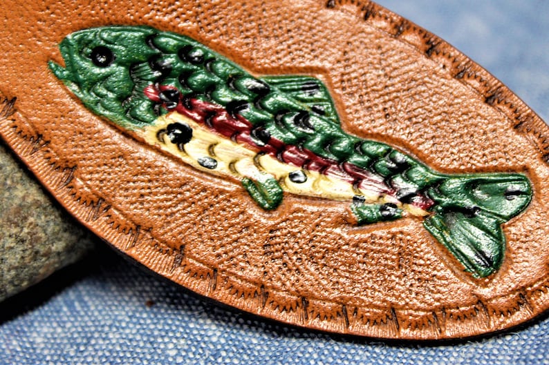 May include: A brown leather oval with a detailed, painted trout design. The fish is green, red, and yellow, with black accents. The leather has a textured surface and a stitched border, suggesting a handcrafted item.