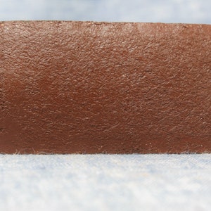 Puede incluir: Una etiqueta rectangular de cuero marr&oacute;n para llavero con una superficie texturizada. La etiqueta tiene una esquina redondeada y un anillo de metal para sujetar las llaves. El fondo es una tela azul claro.