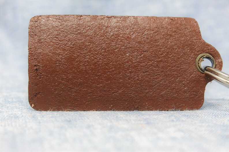 Puede incluir: Una etiqueta rectangular de cuero marr&oacute;n para llavero con una superficie texturizada. La etiqueta tiene una esquina redondeada y un anillo de metal para sujetar las llaves. El fondo es una tela azul claro.
