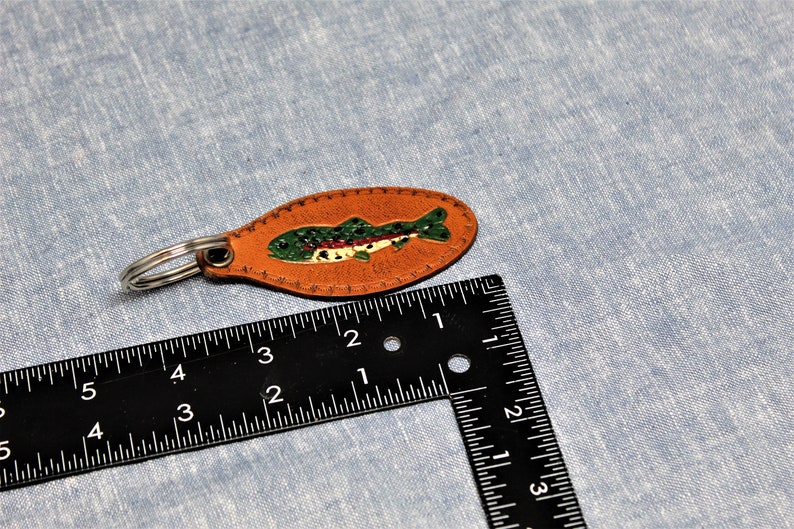 May include: A brown leather keychain with a painted fish design. The fish is green with red and white accents. The keychain is approximately 3 inches long.