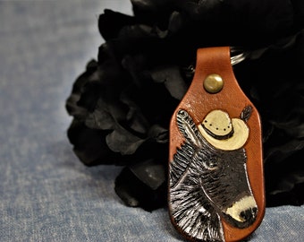 May include: A brown leather keychain featuring a hand-tooled image of a donkey wearing a cowboy hat. The keychain has a brass rivet and a metal ring for keys. The donkey is painted in shades of black, white, and tan.