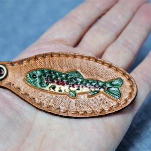 May include: A brown leather keychain with a detailed, colorful fish design. The fish is green, red, and yellow, with a textured appearance. The keychain has a metal ring and a stitched border.