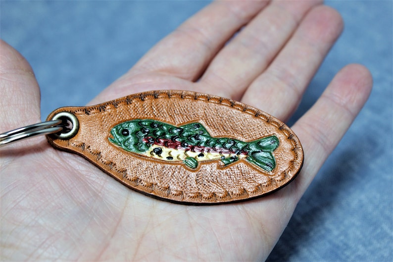 May include: A brown leather keychain with a detailed, colorful fish design. The fish is green, red, and yellow, with a textured appearance. The keychain has a metal ring and a stitched border.