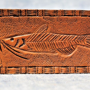 Puede incluir: Un llavero de cuero marr&oacute;n con un dise&ntilde;o de pez tallado. La etiqueta rectangular tiene una superficie texturizada y un borde decorativo. Un anillo de metal est&aacute; unido para las llaves.