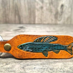 May include: Orange leather keychain featuring a detailed teal fish design. The keychain has a metal ring and a brass rivet. The fish is intricately carved and colored, adding a rustic and outdoorsy feel.