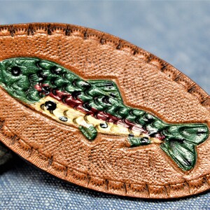 May include: A brown leather keychain with a detailed, hand-painted trout design. The fish is depicted in shades of green, red, and yellow, with a textured surface and stitched border. A metal ring is attached for keys.