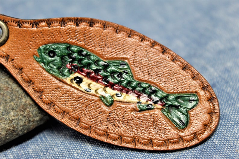 May include: A brown leather keychain with a detailed, hand-painted trout design. The fish is depicted in shades of green, red, and yellow, with a textured surface and stitched border. A metal ring is attached for keys.