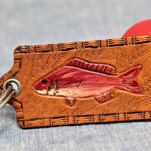 May include: A brown leather keychain with a red fish design. The fish is embossed on the rectangular tag, which has a decorative border. A silver key ring is attached. A red and white bobber is in the background.