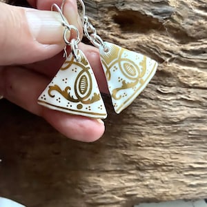 May include: Triangular earrings with a white base and gold decorative patterns. The earrings are suspended from silver hooks. The gold design features swirls and shapes. The earrings are displayed against a wooden backdrop.