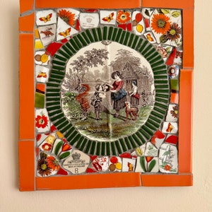 May include: A mosaic artwork featuring a vintage transferware plate depicting a scene of people in a garden. The plate is surrounded by a green and orange mosaic border with floral and butterfly accents. The plate has the text "Royal Standard" and "John Maddock & Sons Ltd. Made in England" on it.