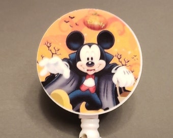 May include: A round Halloween-themed badge reel featuring Mickey Mouse dressed as a vampire. The design includes a pumpkin, bats, and bare trees against an orange background. The reel has a white plastic clip and a metal pin.