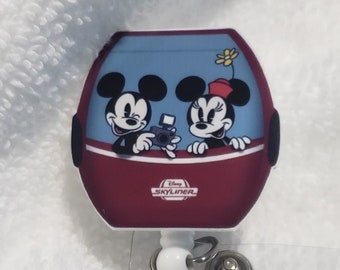 May include: A red and blue Mickey Mouse and Minnie Mouse themed retractable badge holder with a clear strap. The badge holder features Mickey and Minnie in a red and blue ride with the text "Disney's  Hollywood Studios" on the bottom.