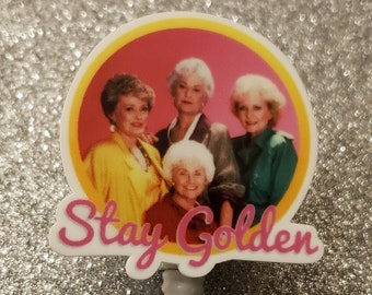 May include: A white plastic badge holder with a round, pink and yellow image of four women. The image has the text "Stay Golden" written below it.