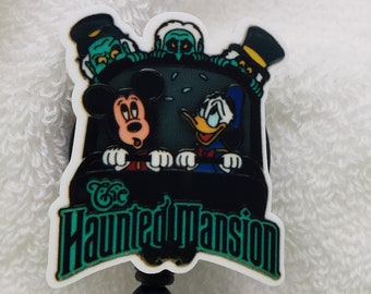 May include: A black and white badge reel with a green and white image of Mickey Mouse, Donald Duck, and two ghosts riding in a doom buggy. The text "The Haunted Mansion" is printed on the badge reel.