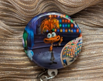 May include: A round button with a cartoon character from the movie Inside Out. The character is orange and has a blue background. The button is attached to a retractable badge holder.
