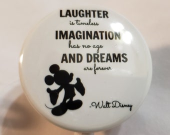 May include: White button with a black silhouette of Mickey Mouse and a quote by Walt Disney: "Laughter is timeless, imagination has no age, and dreams are forever."