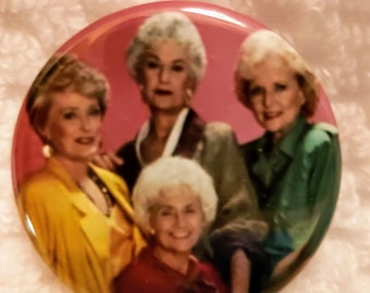May include: A round, glossy button featuring a group portrait of four women. The women are dressed in various outfits, including a yellow blazer and a green blouse. The button has a pink background and is likely a collectible item.