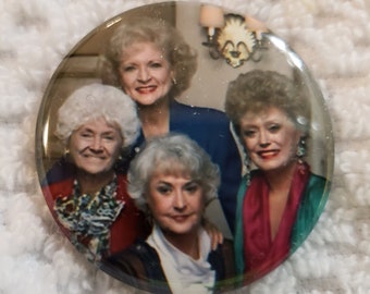 May include: A round button featuring a photo of four women with white hair. The women are smiling and wearing colorful clothing. The button is clear and has a glossy finish.